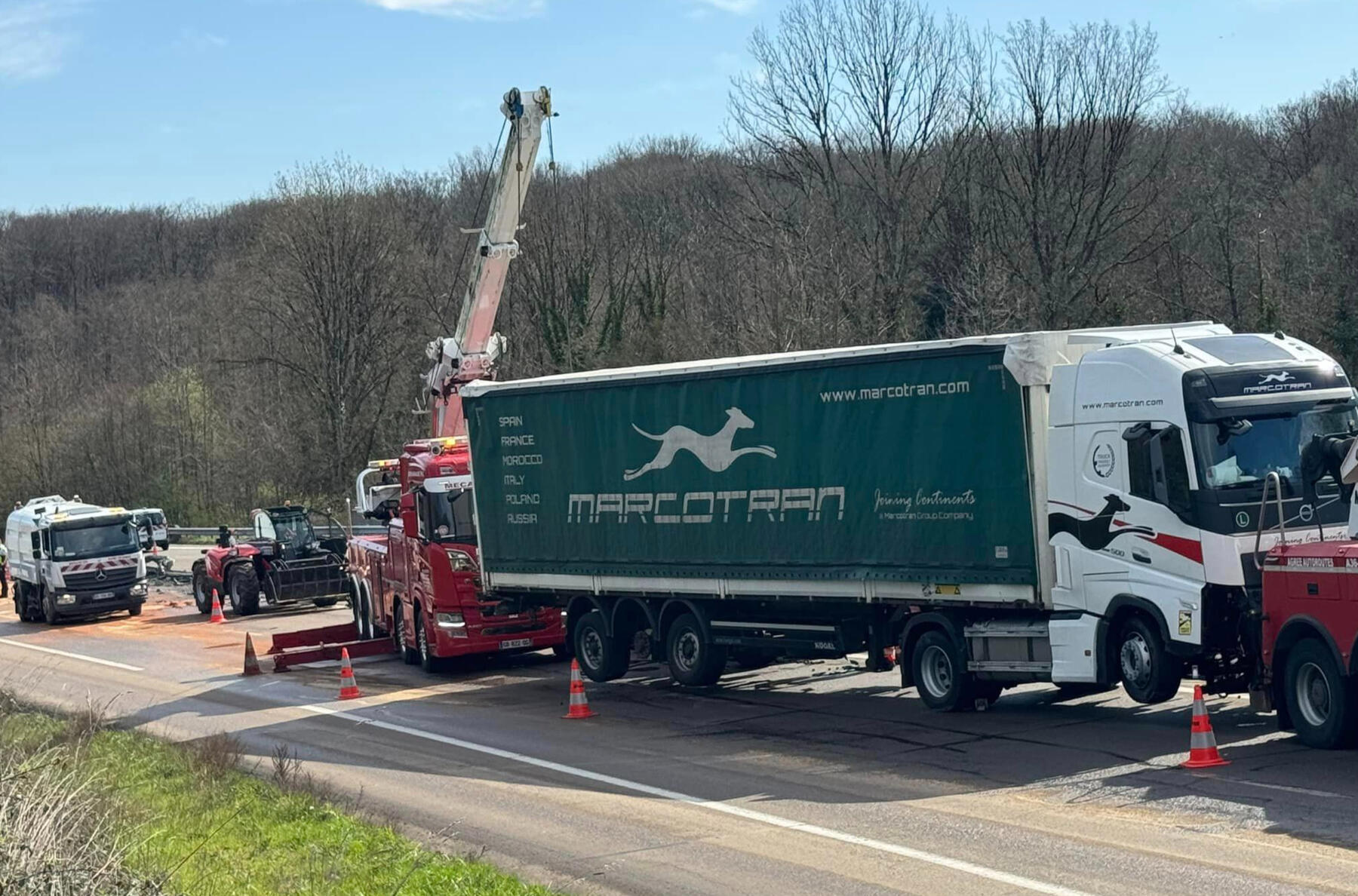 Relevage Poids Lourd Accidenté | Intervention spécialisée Mecano Service FC sur A36 axe de Besançon Intervention d'urgence de Mecano Service FC sur l'autoroute A36 près de Besançon. Deux dépanneuses poids lourds rouges avec grue relèvent un camion semi-remorque Marcotran accidenté. Zone sécurisée avec cônes routiers et présence de plusieurs véhicules d'a